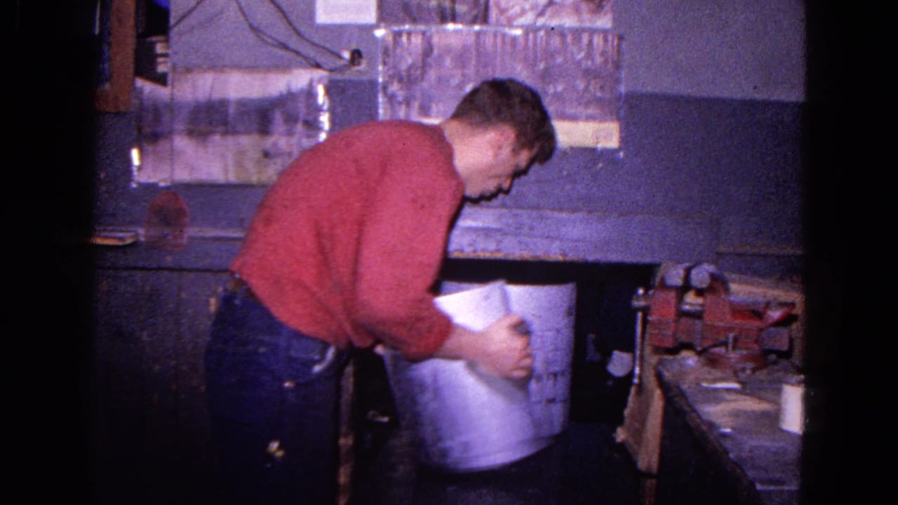 A man in a red sweater and jeans handles a large metal cylinder in a Trenton, New Jersey workshop, with tools visible nearby.