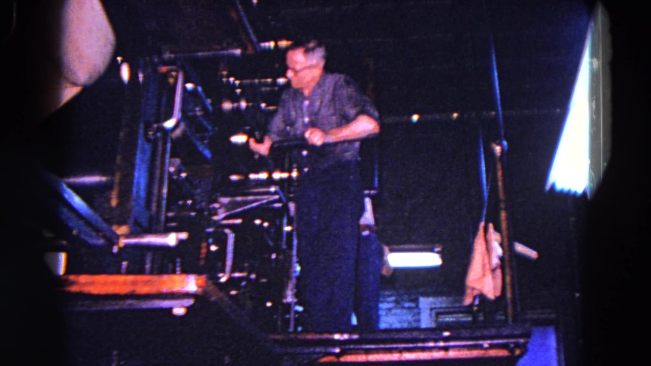 A worker in a dark shirt stands at an industrial machine in a dim Trenton workshop or factory scene from the 1967 home movie.
