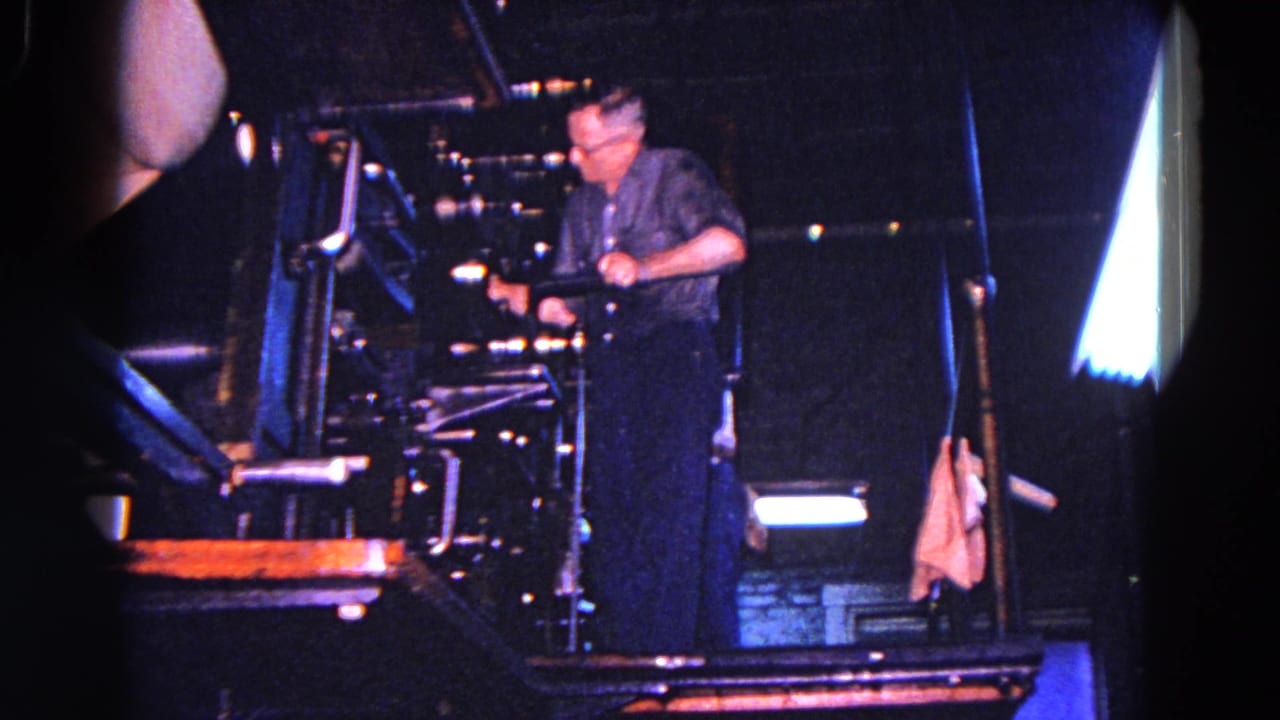 In 1967 Trenton, New Jersey footage, a man in glasses stands at a large industrial printing press in a dim workspace.