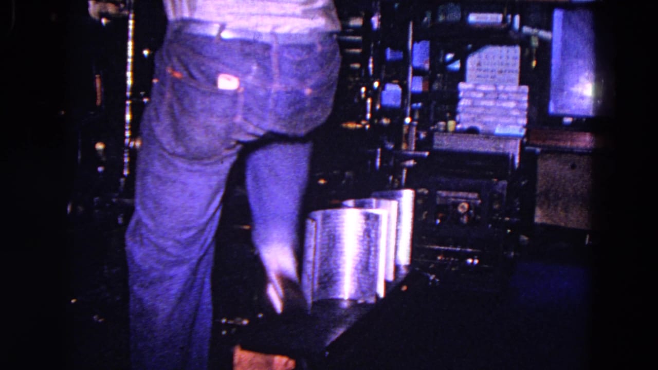 A closer low view shows the worker's legs near the press, with stacked paper or printed material and machinery in the factory interior.