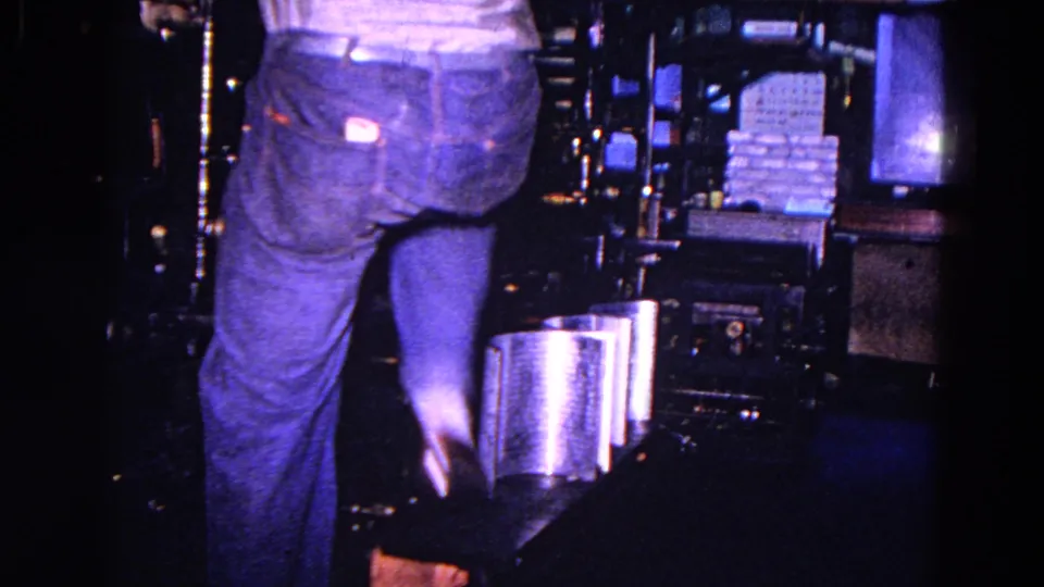 A closer low view shows the worker's legs near the press, with stacked paper or printed material and machinery in the factory interior.