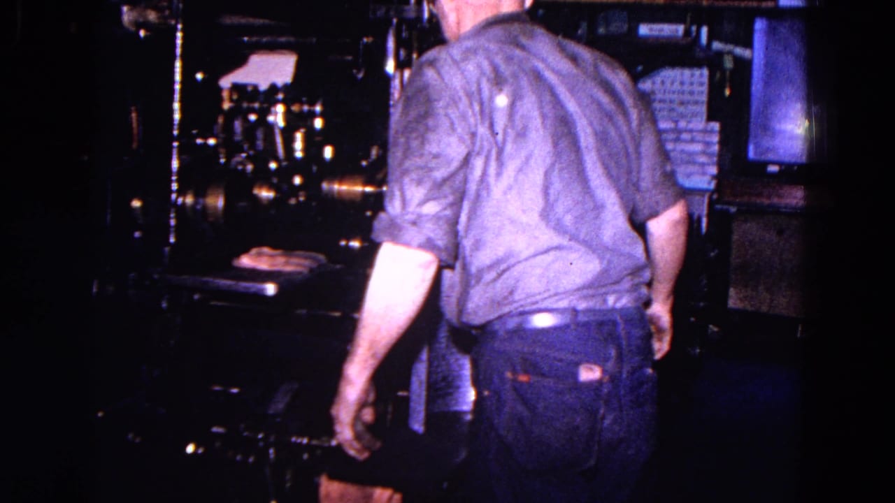 The worker is seen from behind in the 1967 Trenton industrial interior, standing near the press, rollers, and stacked material.