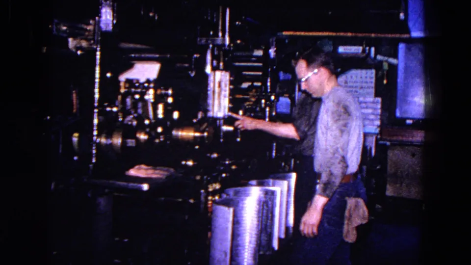 In the Trenton factory scene, a bespectacled worker reaches toward controls or parts on a large industrial machine.