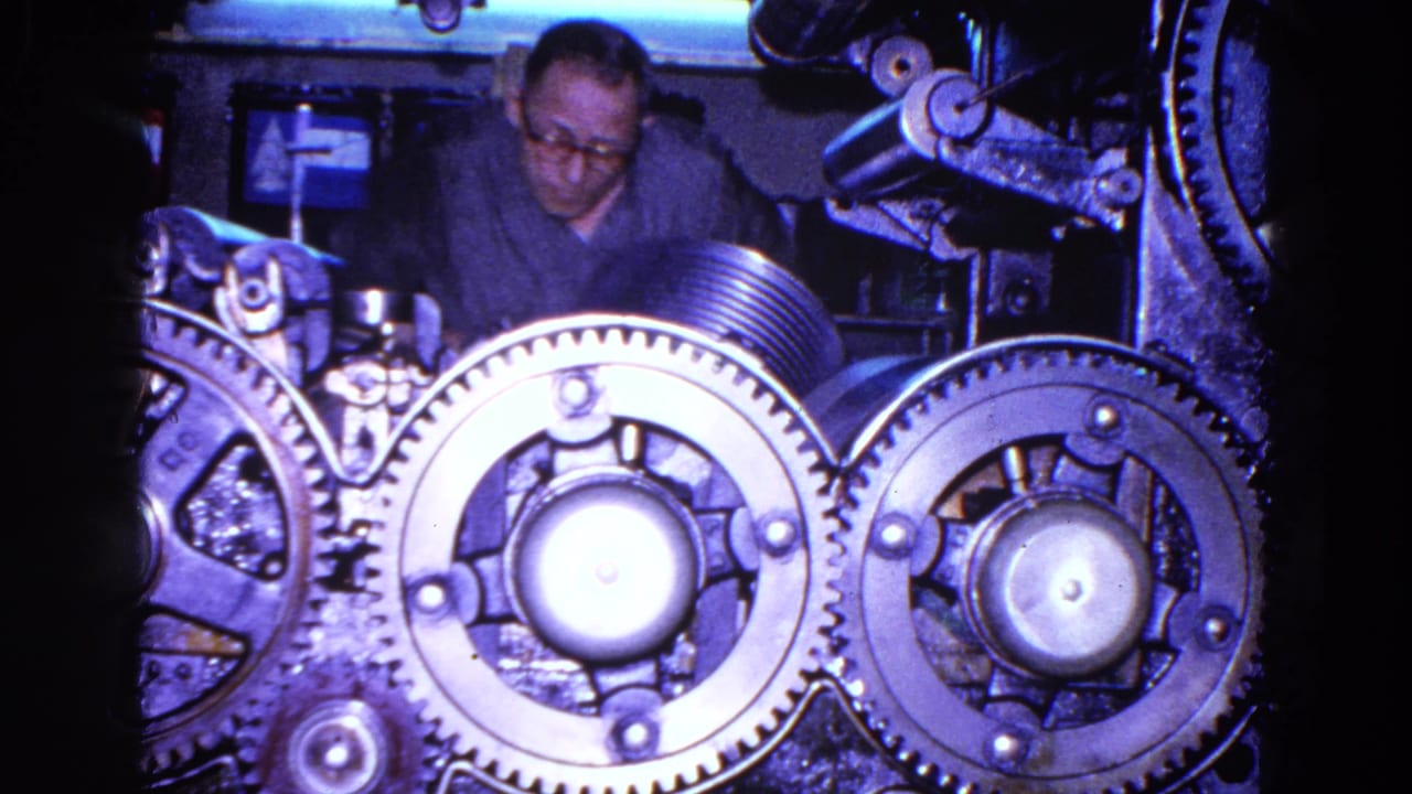 Close-up of large gears and rollers with an adult worker visible behind the machinery in the 1967 Trenton factory clip.