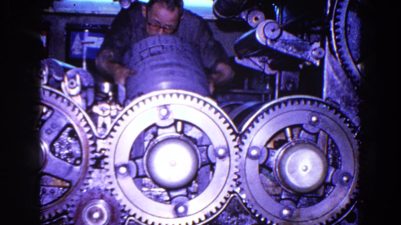 Man with glasses leans over a large industrial machine with toothed gears.