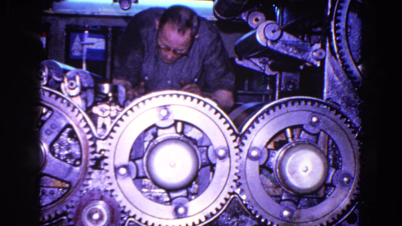 The man leans forward over the industrial mechanism as bright metal gears dominate the 8mm frame.