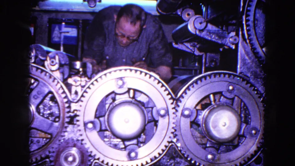 The man leans forward over the industrial mechanism as bright metal gears dominate the 8mm frame.