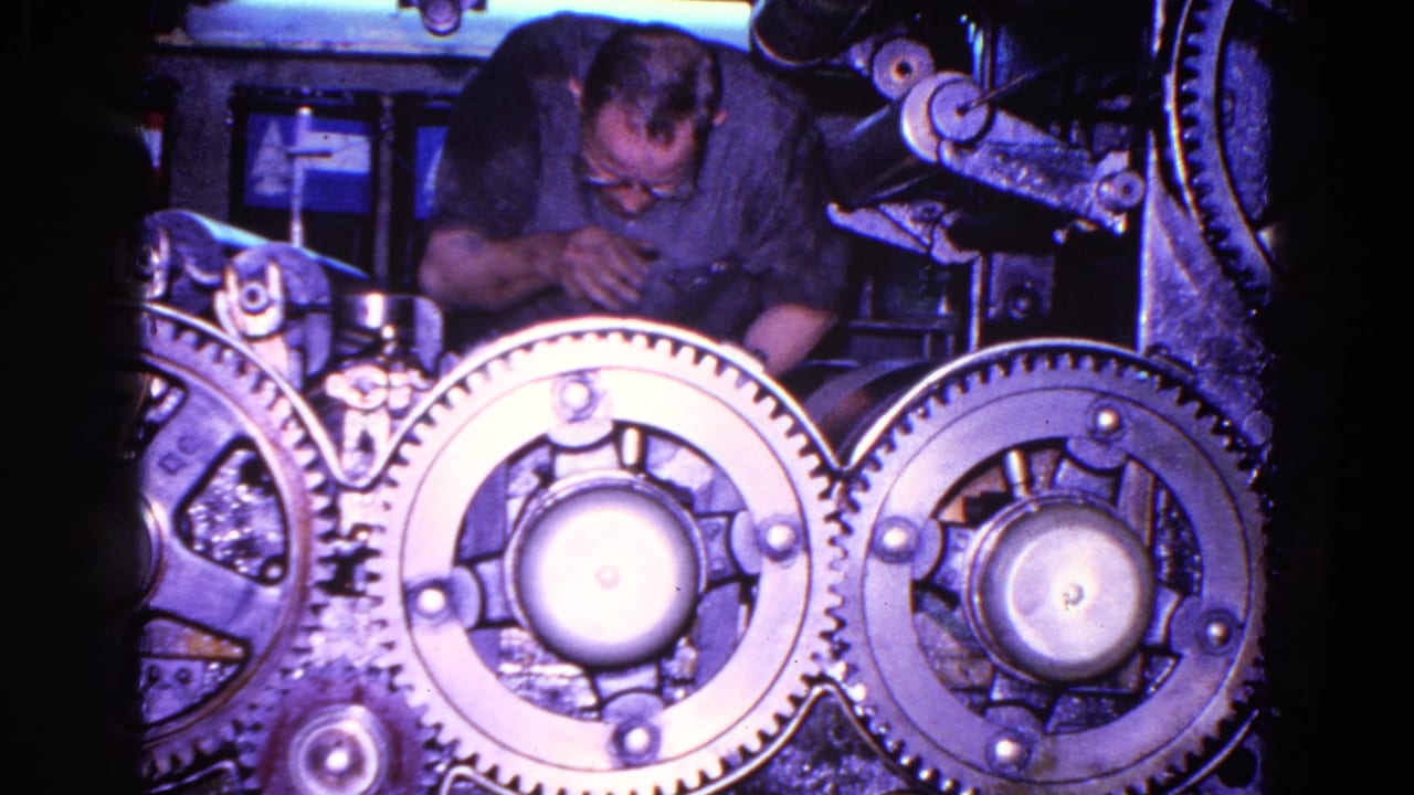 The worker appears to inspect or adjust the machinery, with overlapping toothed wheels and dark mechanical parts in front of him.
