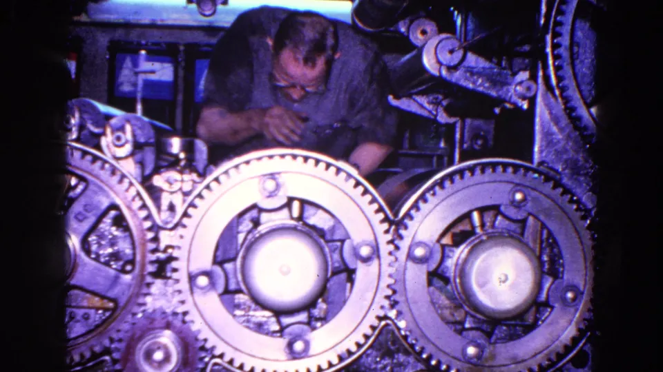 The worker appears to inspect or adjust the machinery, with overlapping toothed wheels and dark mechanical parts in front of him.
