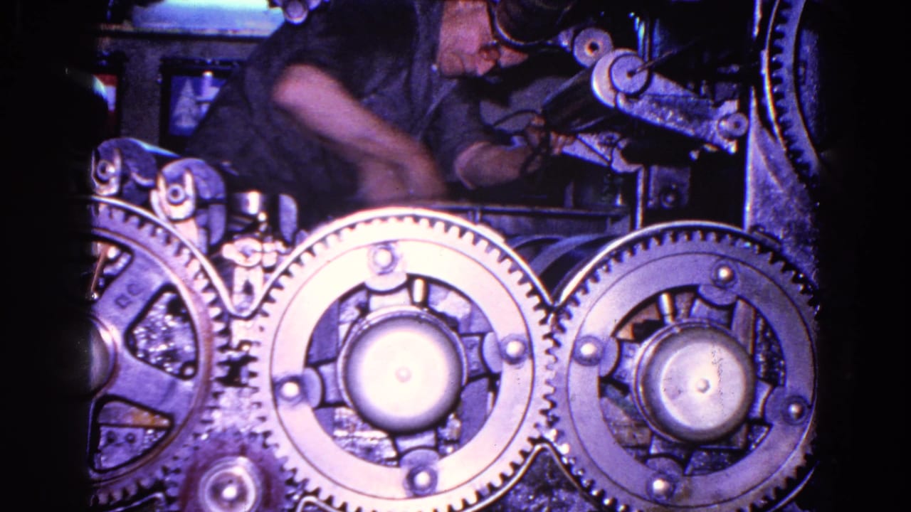 A worker's arm moves across the machine area, with heavy circular gears and metal housings visible under cool, dim light.