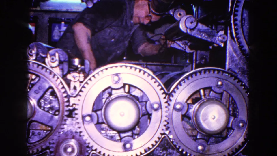 1967 Trenton factory footage showing large exposed gears and part of a worker operating machinery in dim light.