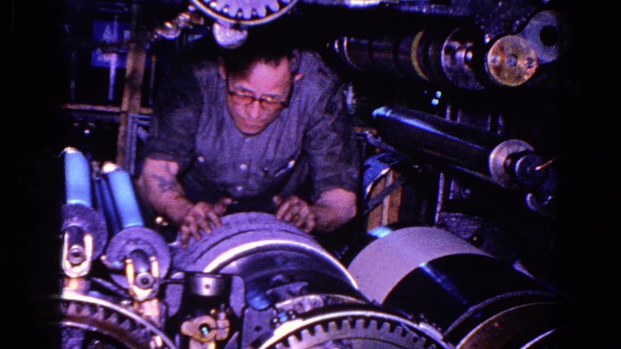 A worker wearing glasses leans over a ribbed cylinder amid gears and rollers in 1967 Trenton factory footage.
