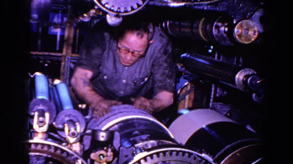 In 1967 Trenton, New Jersey footage, a man in glasses works over exposed gears and rollers on a large industrial machine.