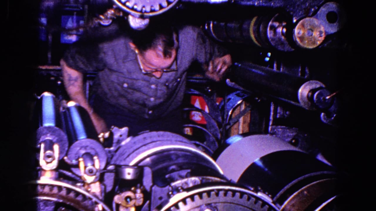 The 8mm frame shows the mechanic leaning into the industrial machine, surrounded by gears, rollers, and dark metal components.