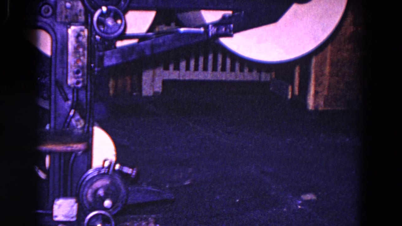 A close 1967 8mm view isolates the industrial machine's rollers, gears, arms, and dark metal surfaces without the worker visible.