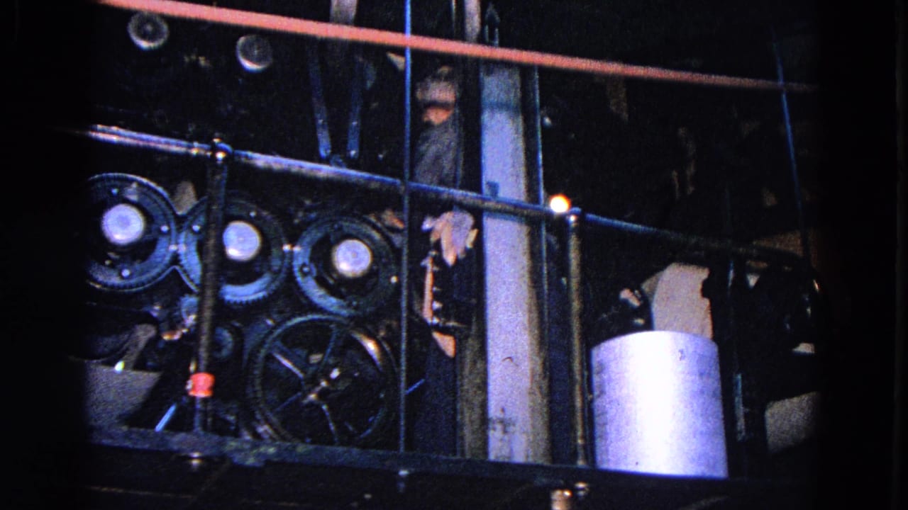 Worker partly visible behind railings beside dark industrial gears and a cylindrical tank.