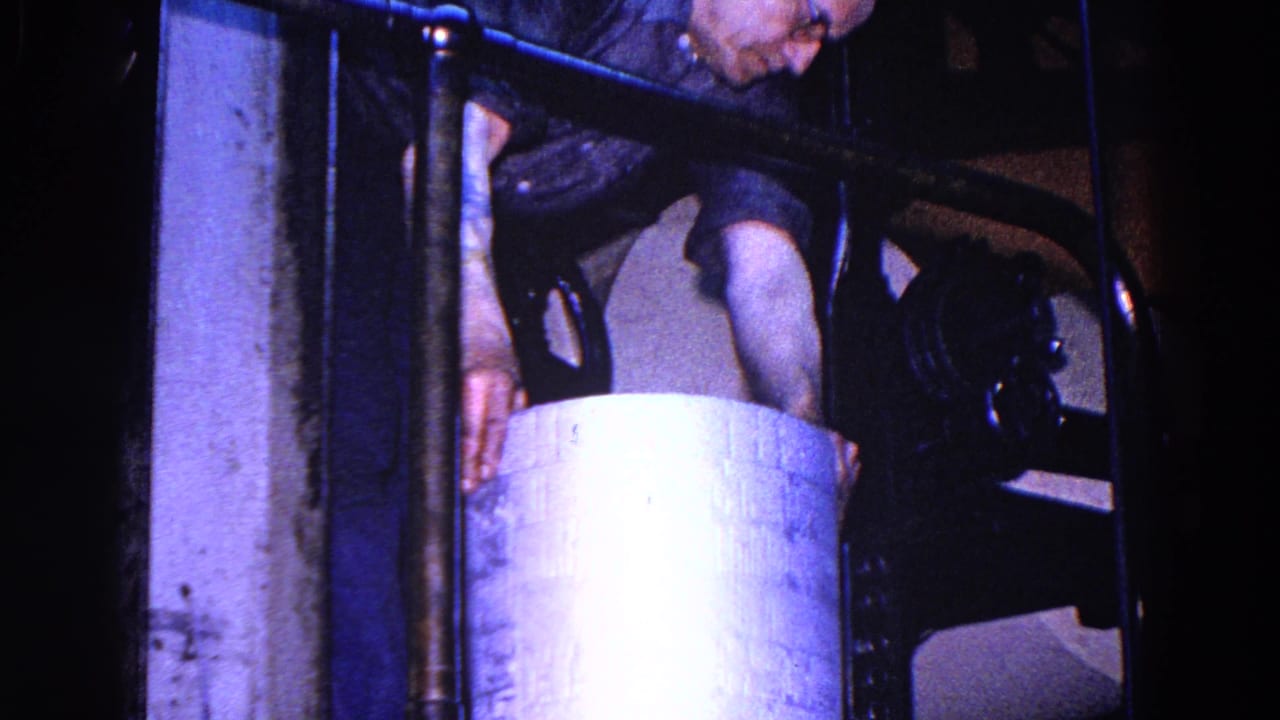 In the Trenton factory footage, a worker bends toward a large cylindrical container beside railings and machinery.