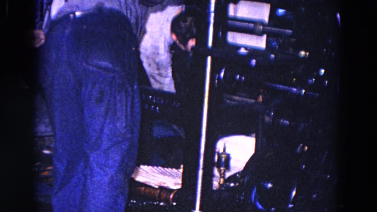 Adult worker bends beside dark paper-handling machinery, with paper sheets and metal components visible in the Trenton factory scene.