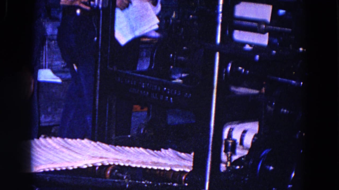 A dim 1967 8mm view from Trenton, New Jersey, shows a worker partly visible near dark printing press machinery and a conveyor carrying sheets of paper.