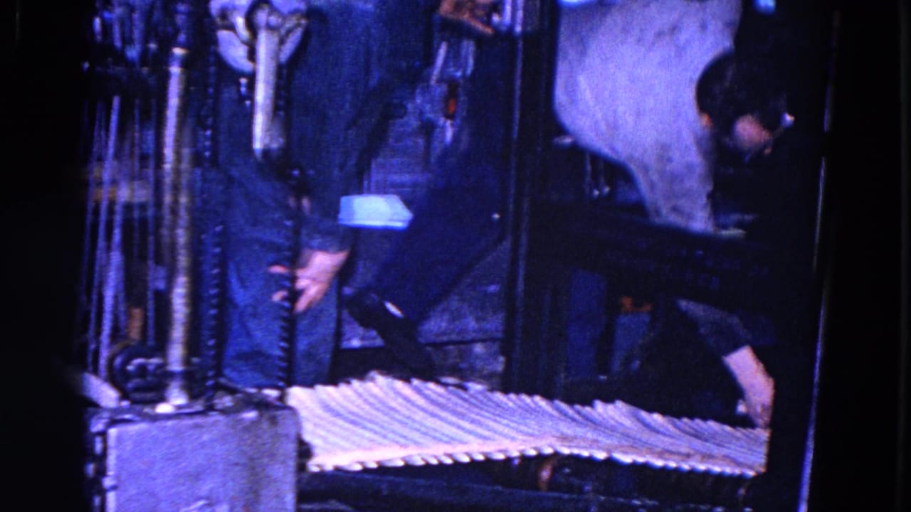 A man in work clothes leans beside a large printing press while a row of paper sheets moves across a conveyor in the dim industrial interior.