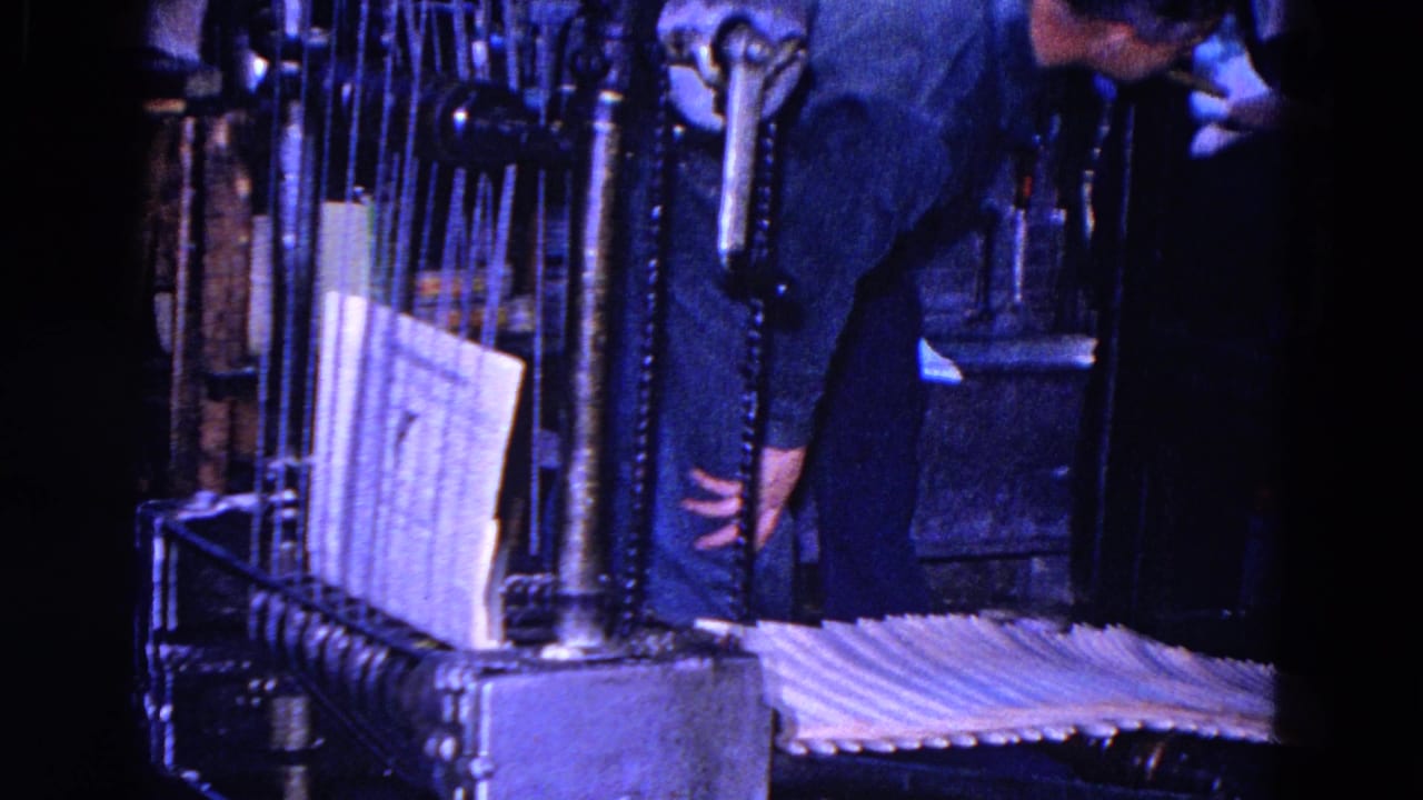 In the blue-tinted 8mm frame, a worker bends over the machinery near a vertical stack of paper and a conveyor section inside the printing area.