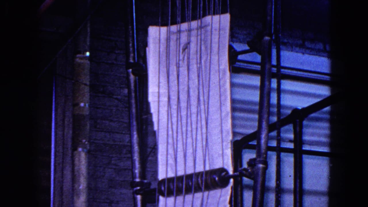 Close view of a printing press area in 1967 Trenton, New Jersey footage, with a pale paper sheet, vertical pipes, a brick wall, and dark machine parts in shadow.
