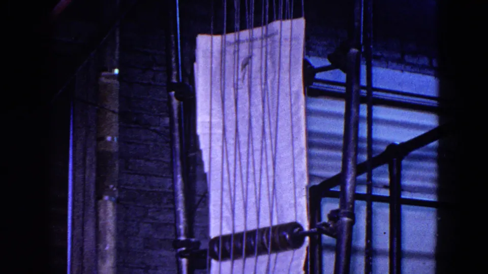 Close view of a printing press area in 1967 Trenton, New Jersey footage, with a pale paper sheet, vertical pipes, a brick wall, and dark machine parts in shadow.