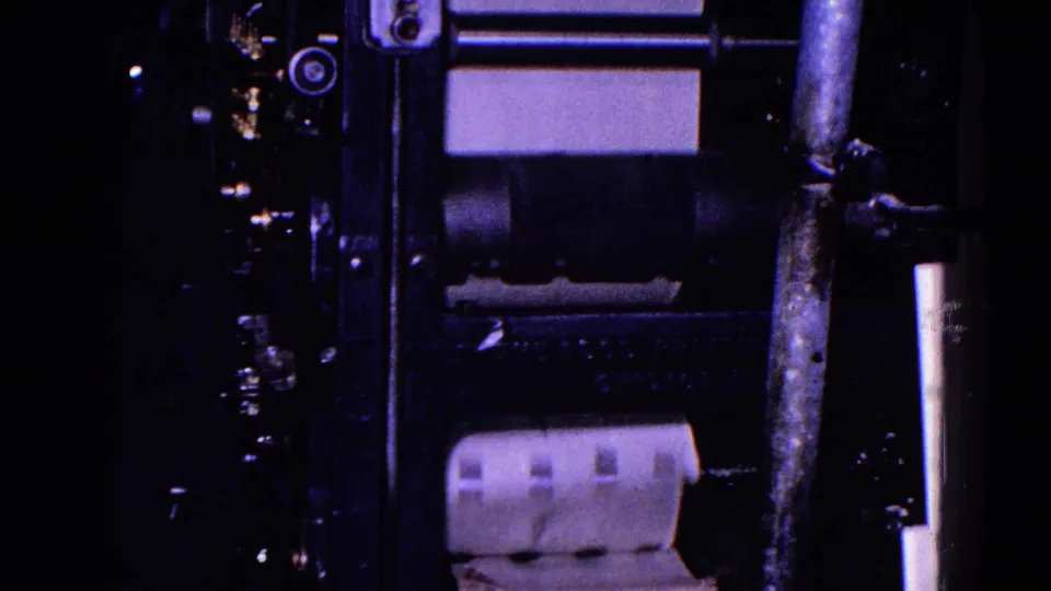 A sheet of paper moves through rollers on a printing press, with metal rods and machine hardware visible in the 1967 Trenton industrial interior.