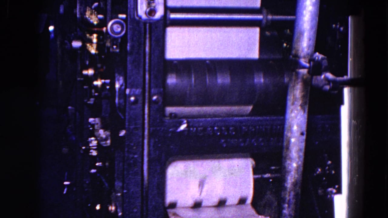 Close-up of a rolling press machine with paper at the lower roller area, vertical piping, and dark metal mechanical components in blue-tinted 8mm footage.