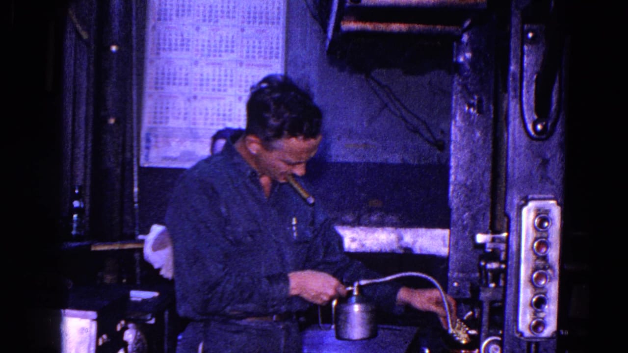 Worker in a blue shirt uses an oil can beside dark industrial machinery.