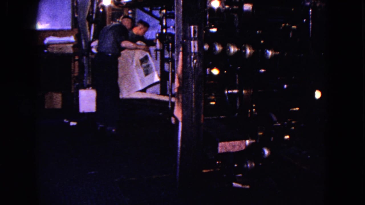 Two workers stand near large machinery while handling a sheet of paper.