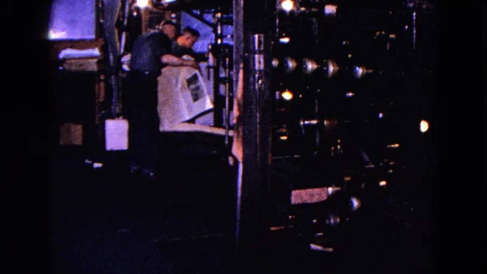Two adult workers appear beside large industrial equipment, with one holding or reading a large paper document in the factory interior.