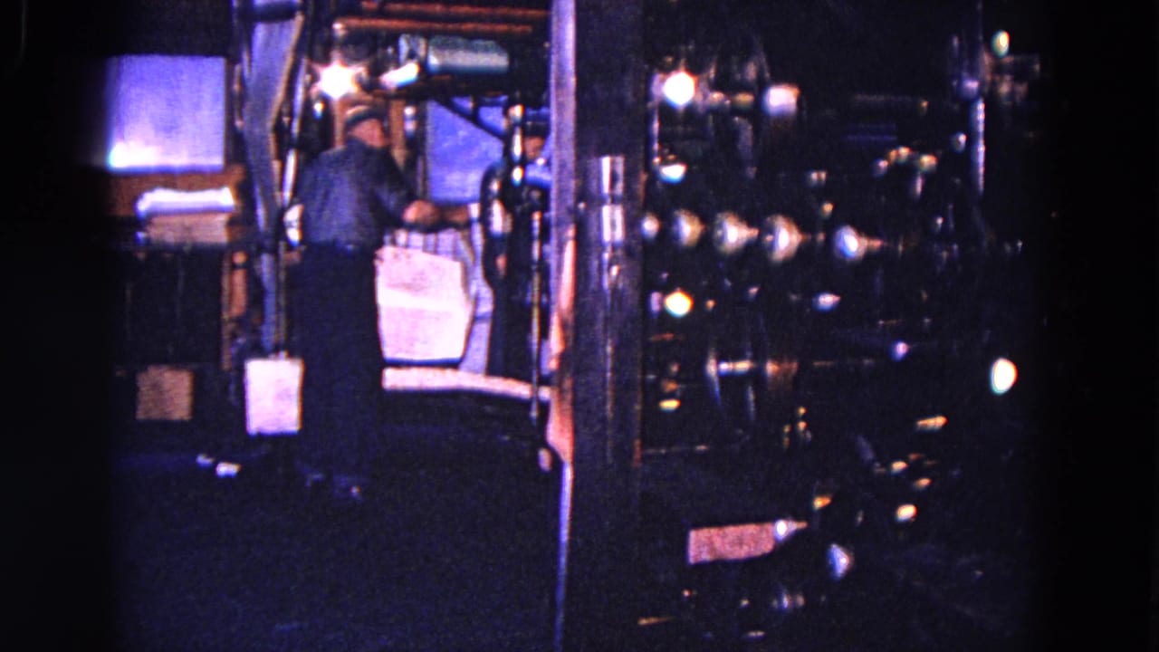 A worker in a blue shirt stands by heavy machinery and holds a large paper sheet, with another worker partly visible nearby.