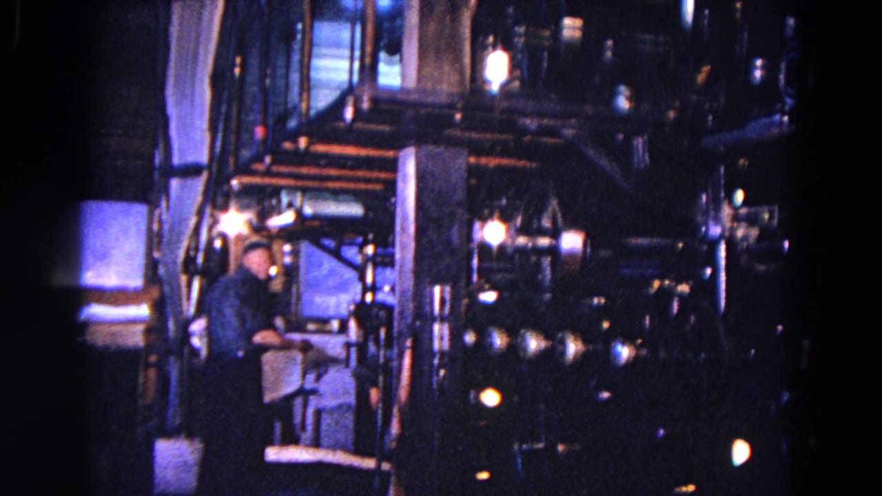 A worker is framed between tall industrial machines inside the 1967 Trenton factory interior, with pipes and rollers visible around him.
