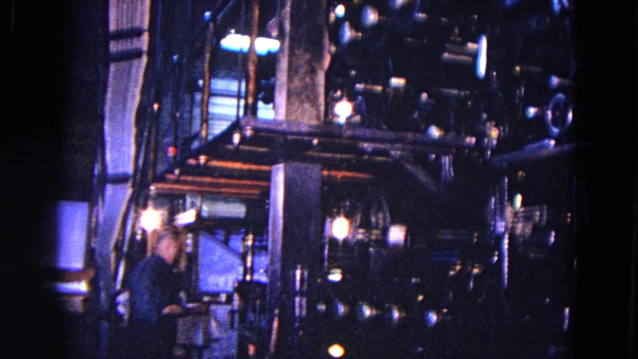 Worker near a dark industrial printing press with metal housings and rollers.