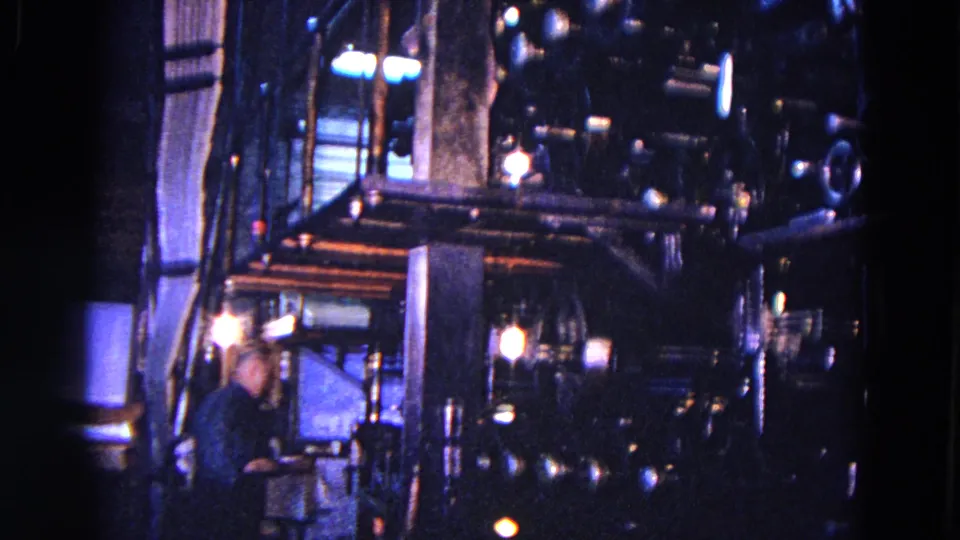1967 Trenton factory footage showing a worker beside a large, dimly lit industrial printing press with exposed machinery and paper handling equipment.