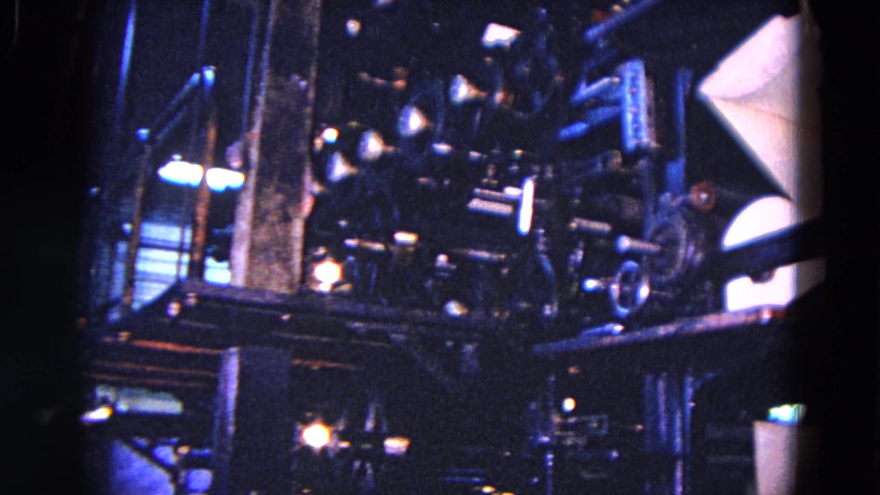 Large printing press mechanisms appear in a dim 1967 Trenton factory view, with rollers, arms, and a pale paper surface at the right edge.