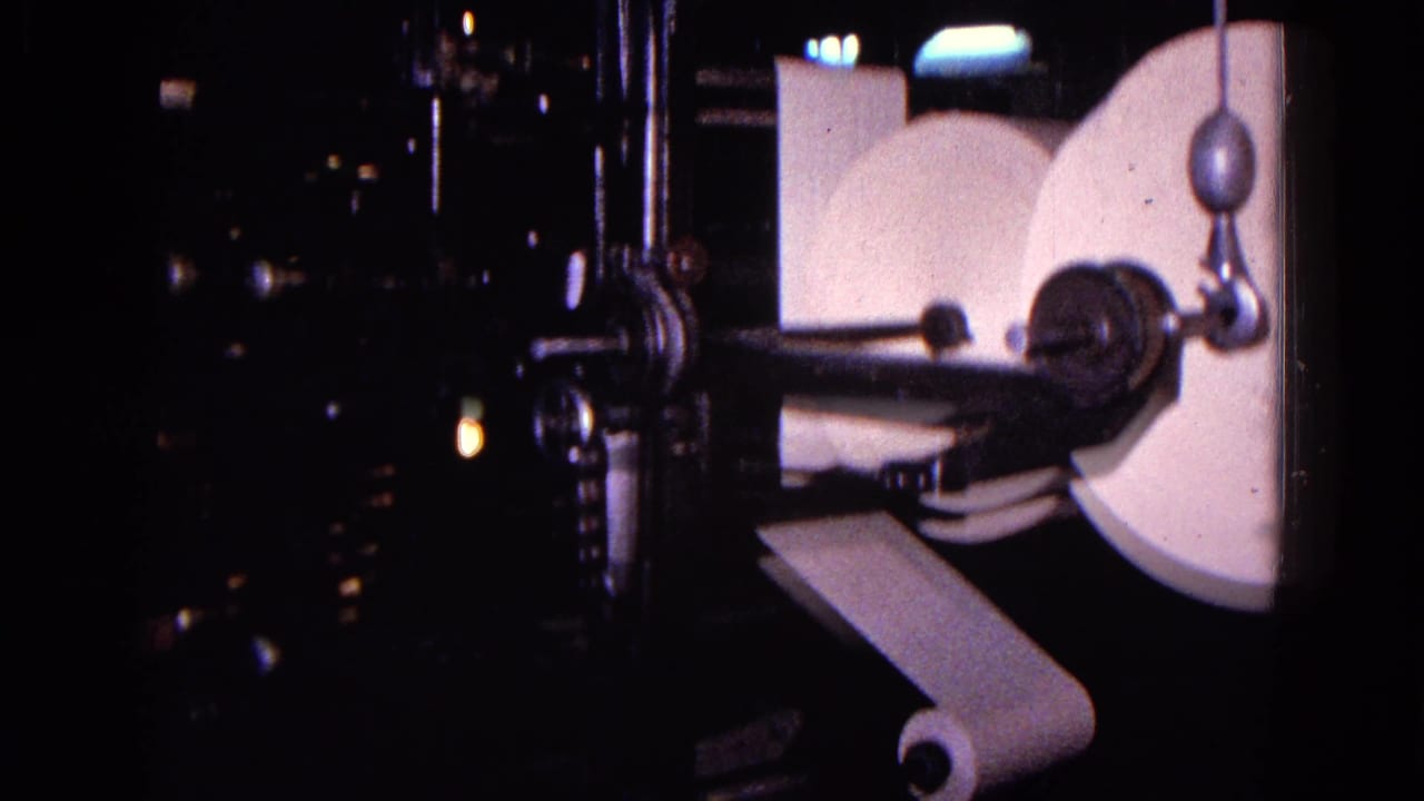 A dim close view of a 1967 Trenton printing press shows large white paper reels, a narrow paper strip, rollers, and dark surrounding machinery.