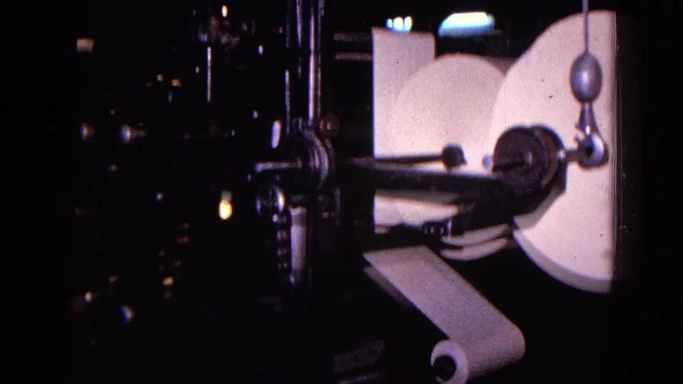 A dim close view of a 1967 Trenton printing press shows large white paper reels, a narrow paper strip, rollers, and dark surrounding machinery.