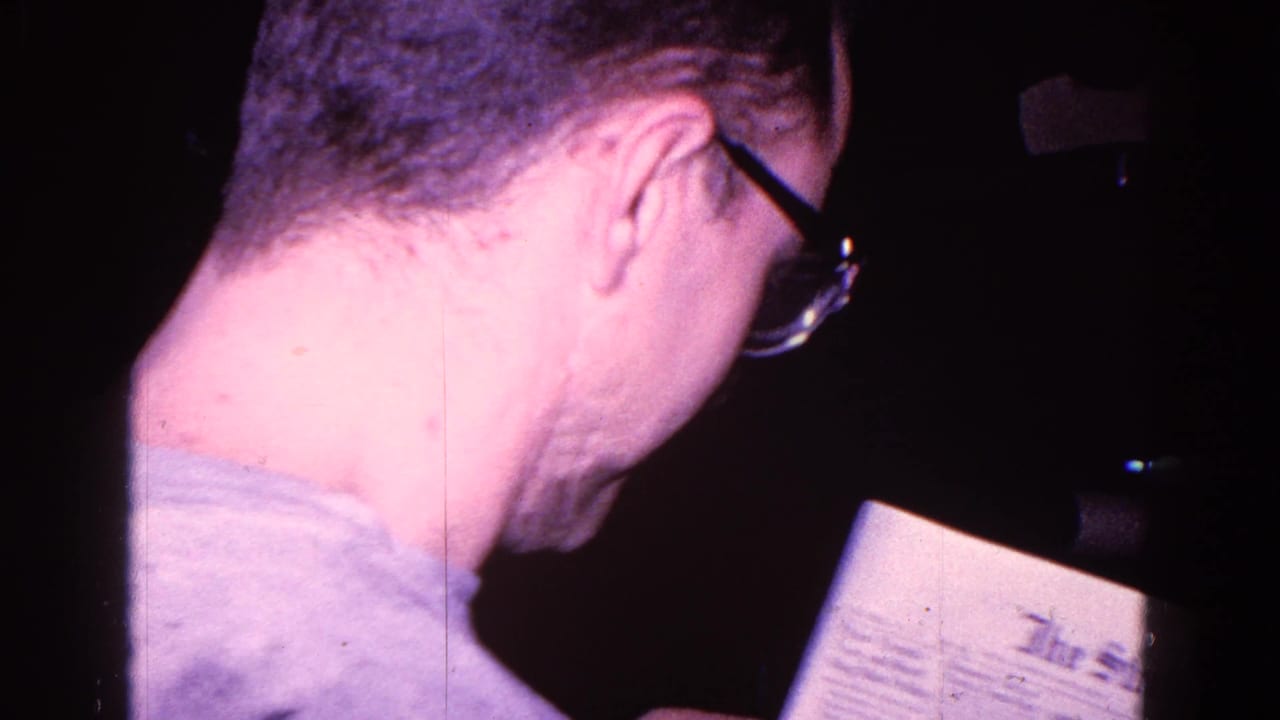 1967 Trenton home-movie frame showing a man in glasses from behind and in profile as he reads a newspaper.