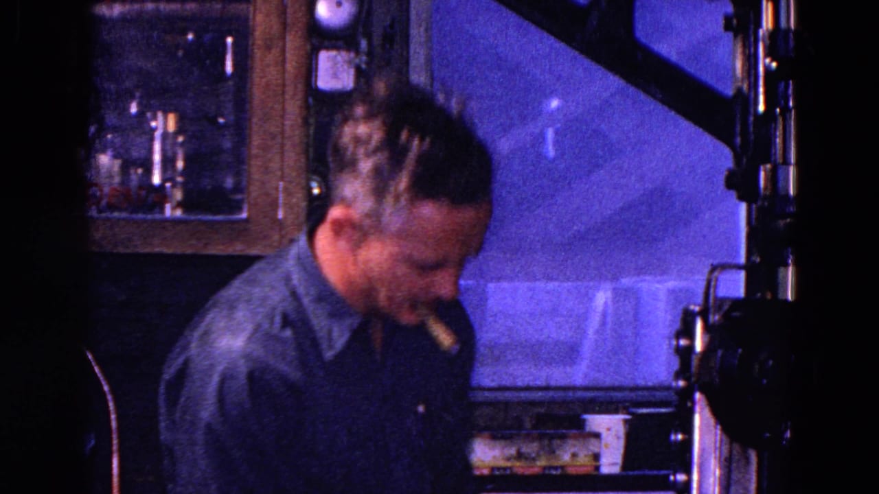 Man with cigar stands beside projection equipment in a dim booth.