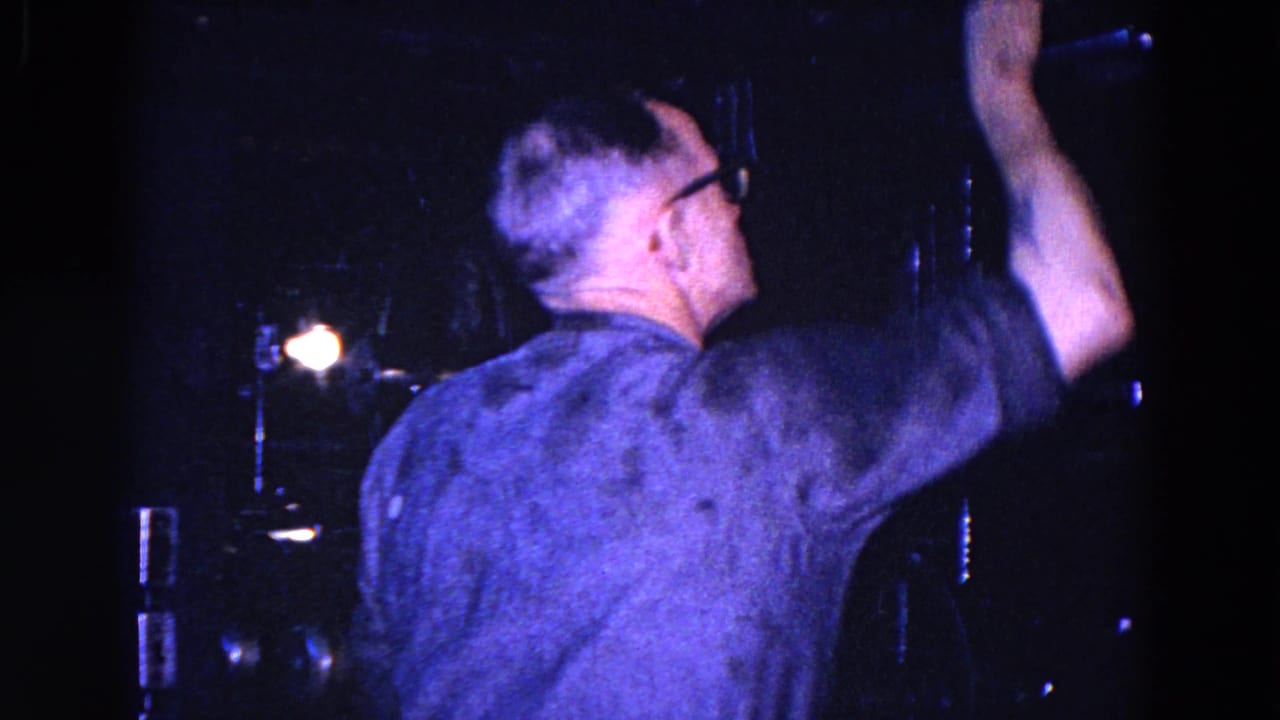 The projection operator, wearing glasses, reaches upward beside dark mechanical equipment in the Trenton booth footage.