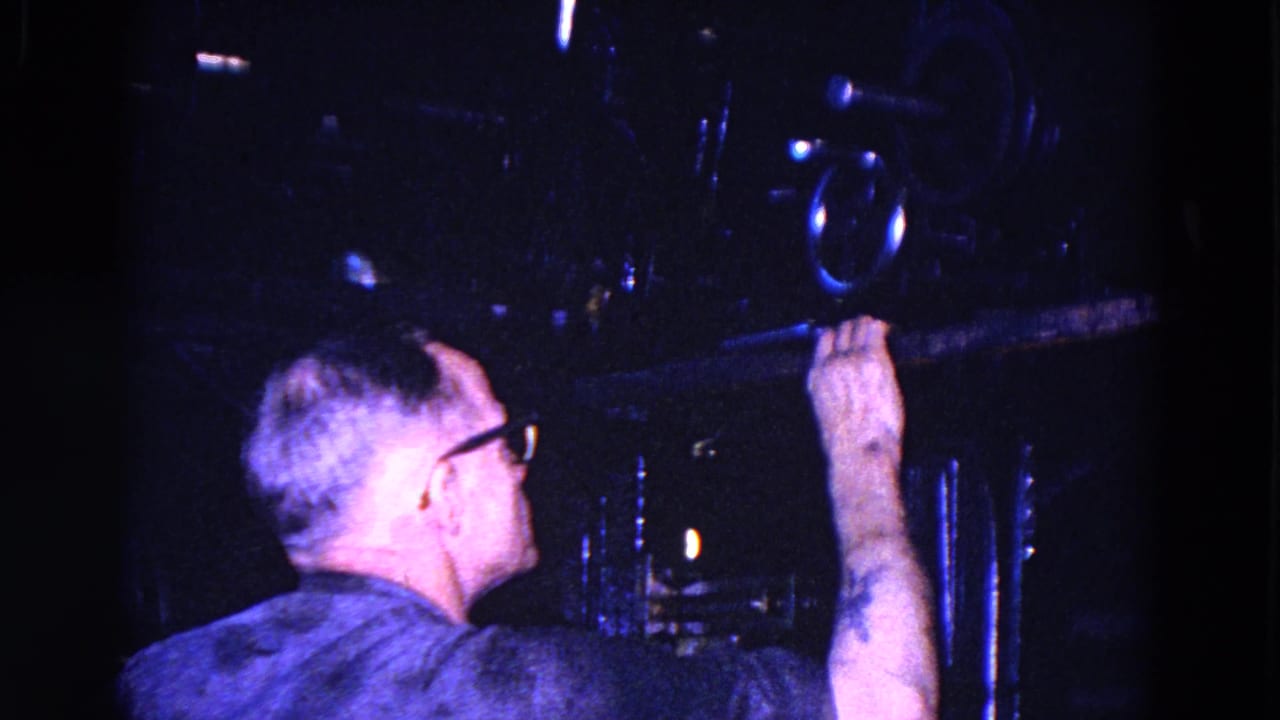A man in glasses handles a horizontal component near film projection machinery in the dim 1967 Trenton booth.
