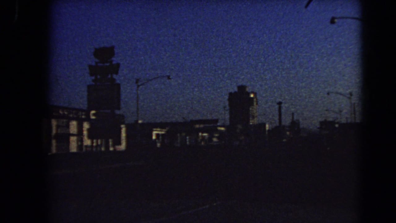 1966 Las Vegas home-movie frame looking down a dark road at dusk toward silhouetted commercial buildings, streetlights, and an illuminated rooftop sign shape.