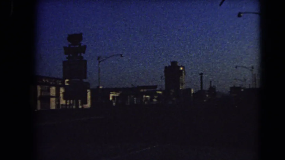 1966 Las Vegas home-movie frame looking down a dark road at dusk toward silhouetted commercial buildings, streetlights, and an illuminated rooftop sign shape.