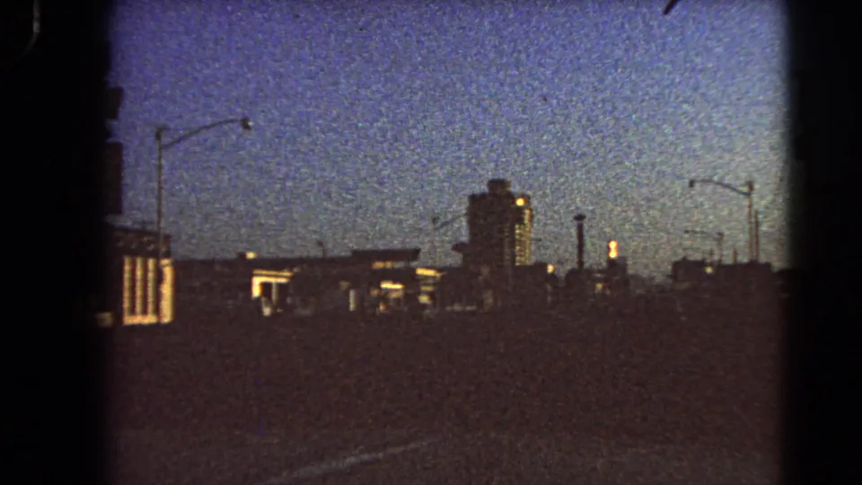 1966 Las Vegas 8mm frame showing a forward road view at dusk with distant lit building fronts, tall streetlights, and dark skyline forms.