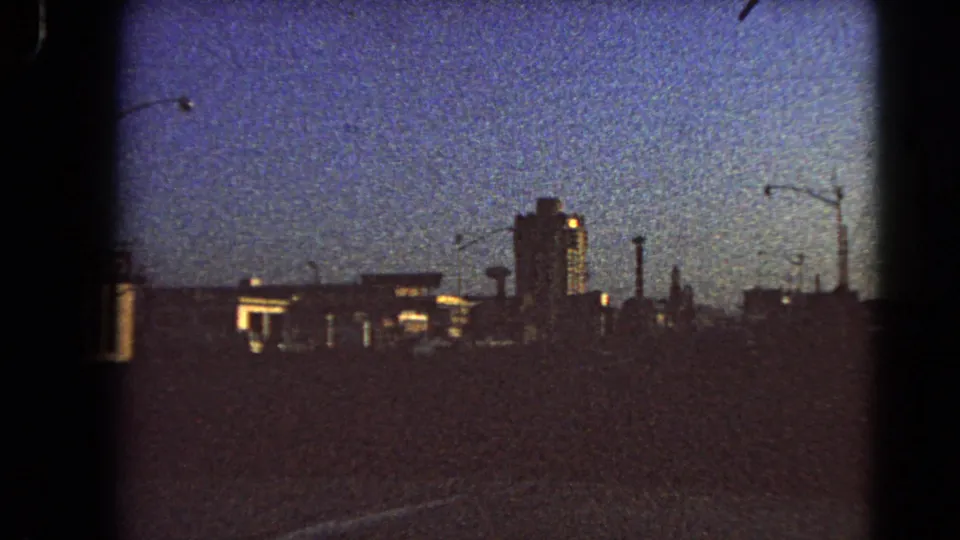 1966 Las Vegas home-movie frame from a slow drive, showing a dark roadway, silhouetted buildings, streetlights, and faint illuminated facades under a blue evening sky.