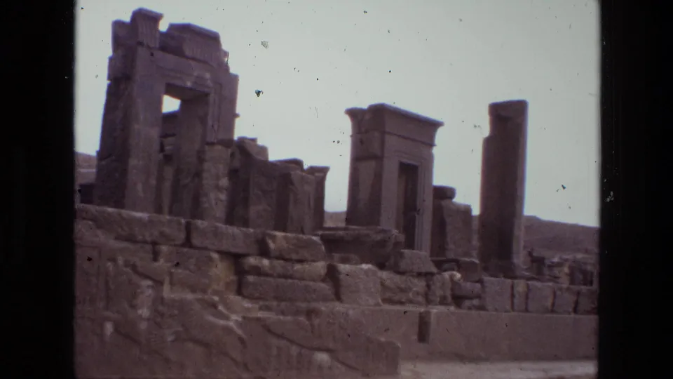 1972 8mm Iran travel frame with block masonry, a tall freestanding column, carved relief stonework, and gateway-like ruins.
