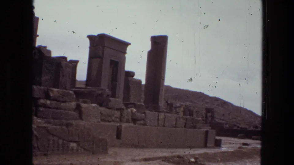 1972 8mm Iran frame showing a doorway-like ruin, upright column, carved stone wall, barren hillside, and dusty foreground.