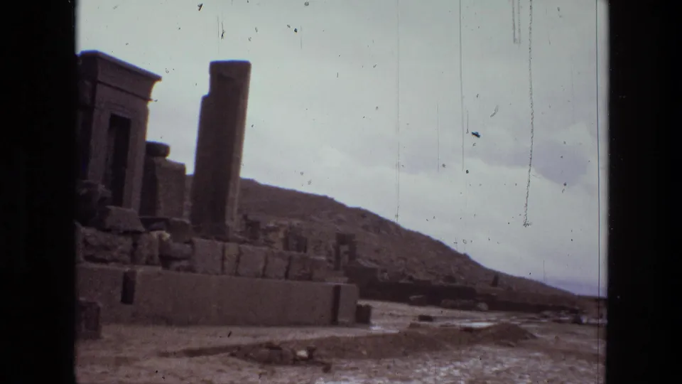 1972 8mm Iran footage showing a stone doorway, freestanding column, low ruin walls, pale sky, and arid hills beyond.